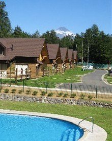 Condominio de cabañas de madera junto a piscina, con volcán nevado de fondo.